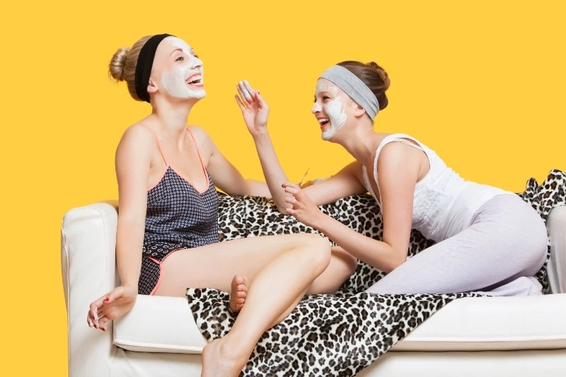 Two women practicing self care using face masks and laughing while sitting on a sofa