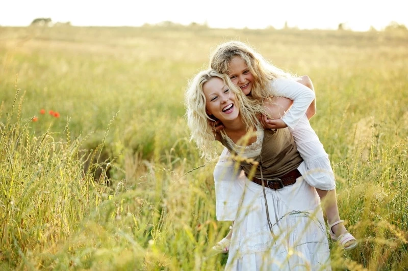 Mother wearing white dress with daughter in a white dress on her back laughing in a field