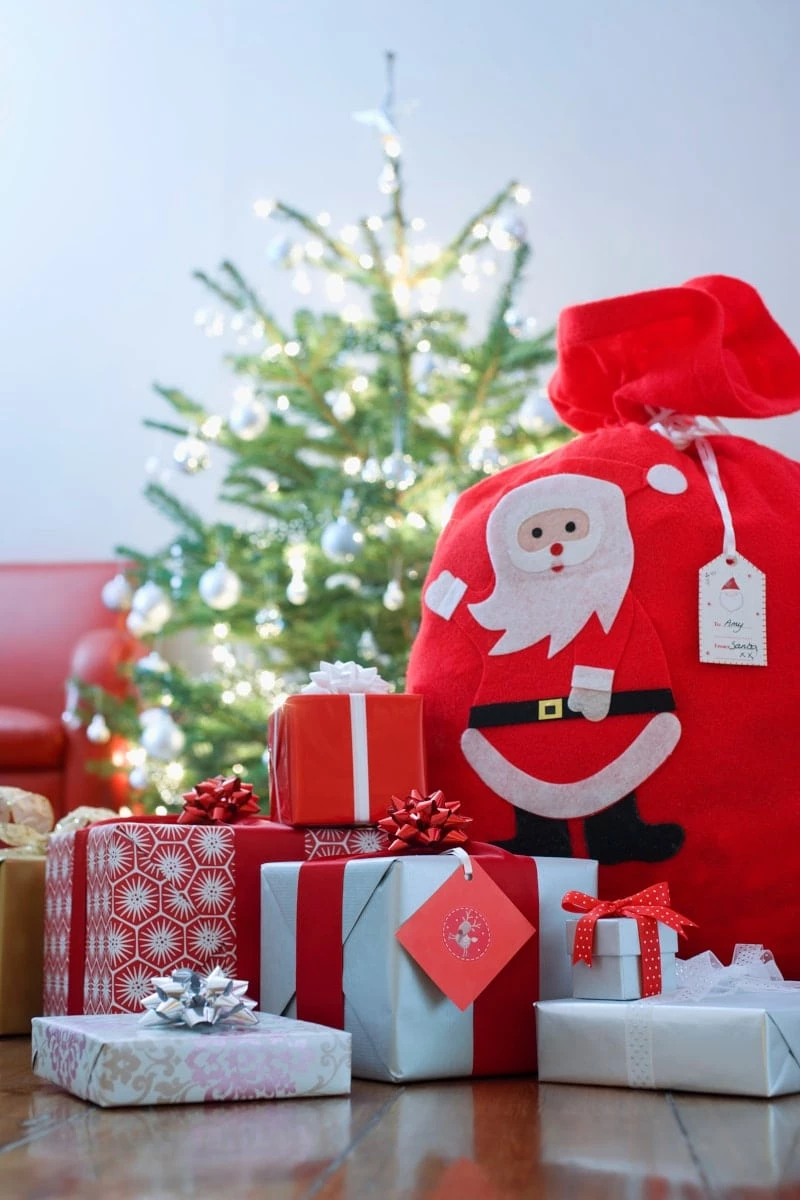 Pile of gifts and a bag with a picture of Santa on it in front of a Christmas Tree