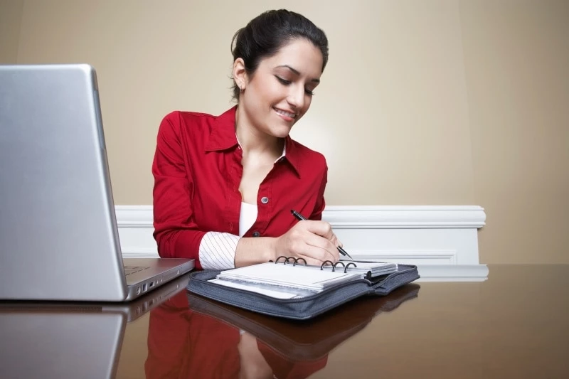 Woman in red shirt writing in a planner while sitting at a laptop