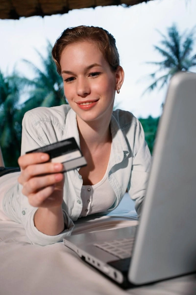 Woman wearing white shirt looking at credit card while sitting at a laptop