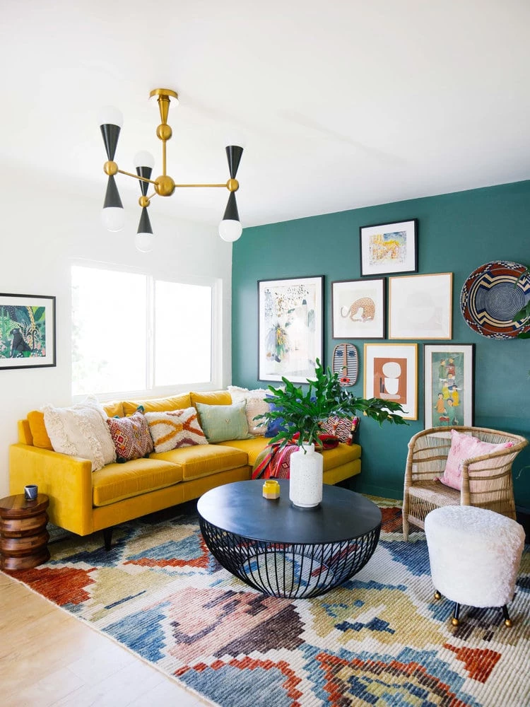 Boho Chic Styled Living Room with a yellow sofa, brightly colored rug, a black circular table, a wicker chair, and a gallery wall on a teal wall.