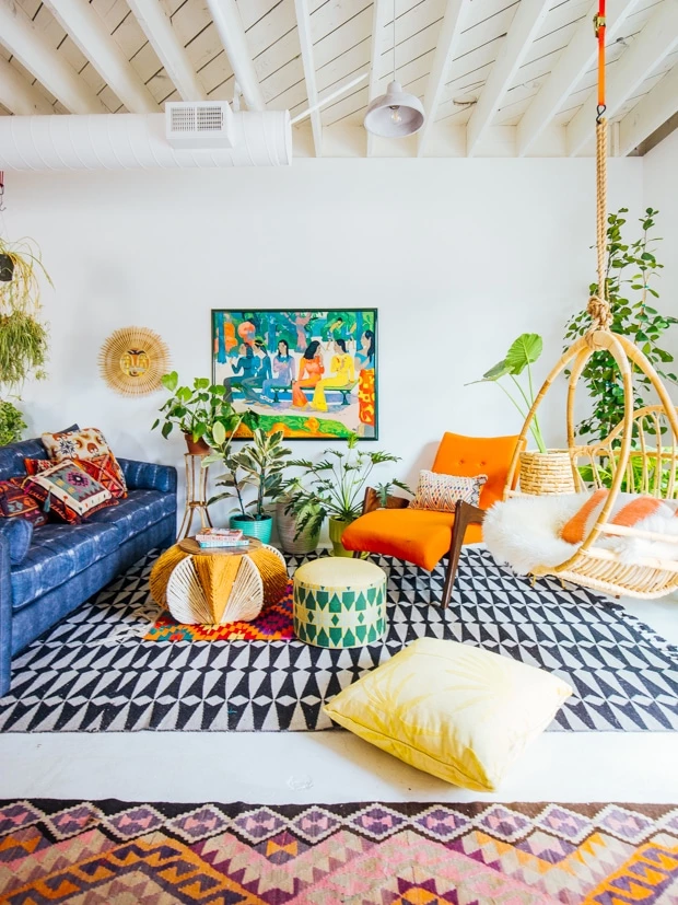 A bold boho chic styled living room with lots of sharp geometric patterns in a rug and pillows. Lots of colors are used in the accessories all over this room with a blue sofa and an orange lounger.