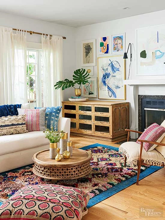 A boho chic styled living room with a a white sofa and a white chair with a wooden frame. A round coffee table that has concrete and gold vases. Lots of blues and pinks in fabrics and artwork in the room.