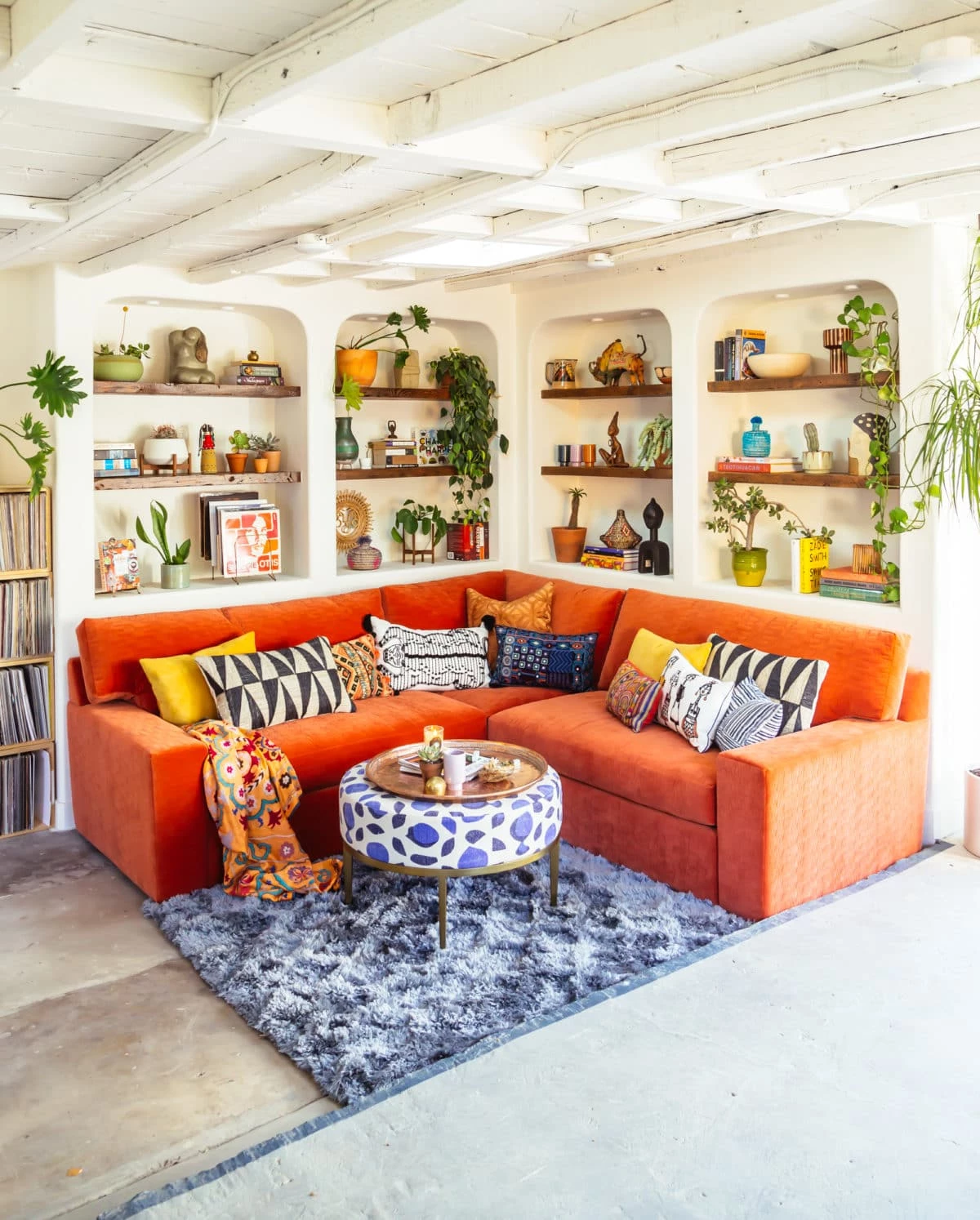 A bright burnt orange sofa with a blue fuzzy rug and bohemian patterns in pillows and a unique coffee table with blue spots. Living room has built in shelves that have lots of books, plants, pictures, and knick knacks displayed.