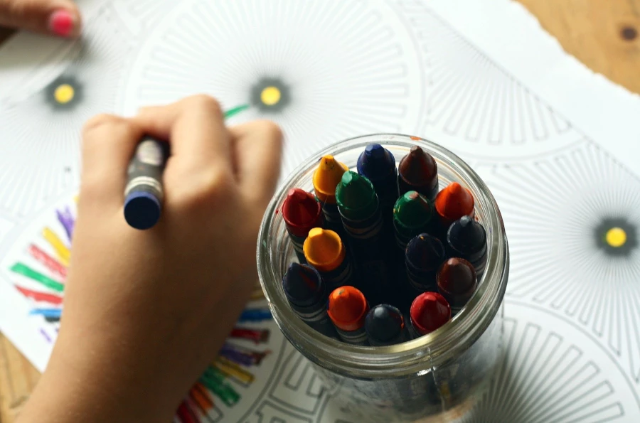 A child's hand coloring with crayons and several crayons in a jar
