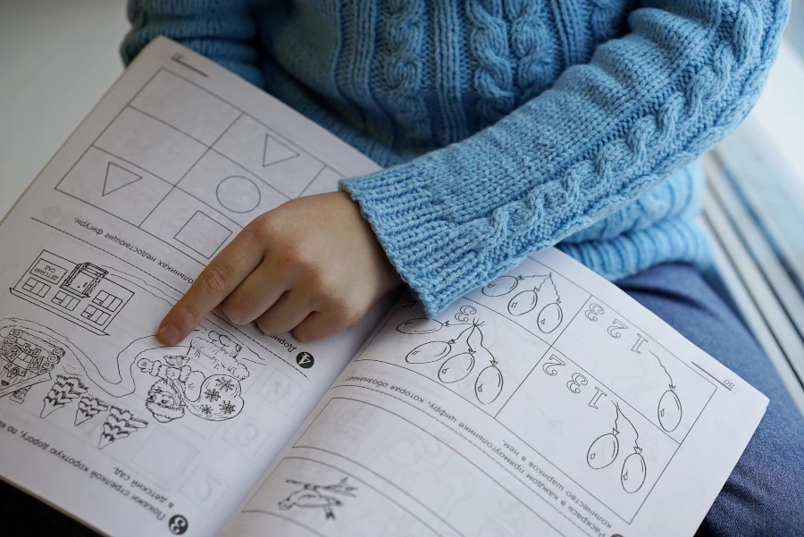Child in a blue sweater pointing to a worksheet in a book